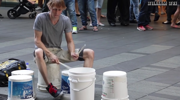 young man holding drumsticks sitting on an overturned bucket, playing other overturned buckets as if they were drums