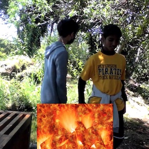 Two people stand outside in a heavily overgrown area with the image of a fire superimposed in front of them