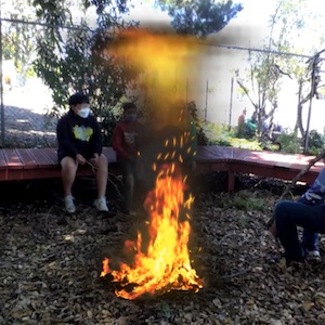 A large campfire burns while three people sit around it