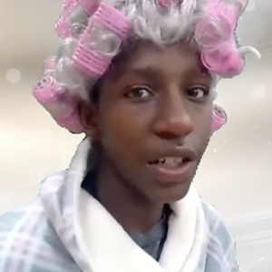 A young man dressed as an old woman has pink curlers in her grey-haired wig