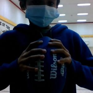A person in a mask standing in a hallway grabs a football with both hands, fingers spread apart.
