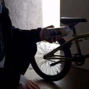 A person kneels near the rear wheel of a bike, a can of spray grease ready to use.