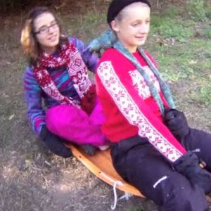 Two people in bright purple and pink winter clothes sit on a sled, ready to move down a hill without snow