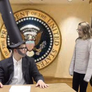 A person sits at a desk with a tall black construction-paper hat with the presidential seal projected on the wall behind him as someone approaches his desk