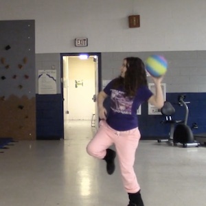 A girl in pink pants and a purple shirt has her arm pulled back with a ball, ready to throw