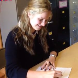 A blonde woman sits at a table smiling while signing some papers