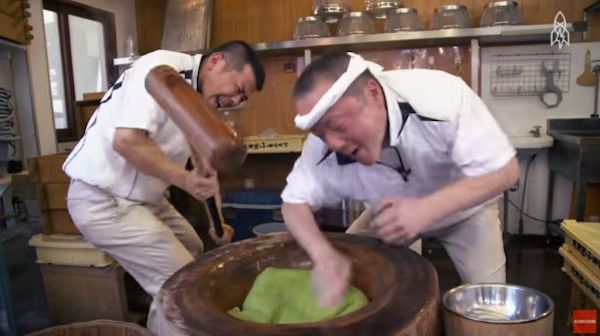 Man leaning over vat with green substance while another man pounds the substance with a hammer