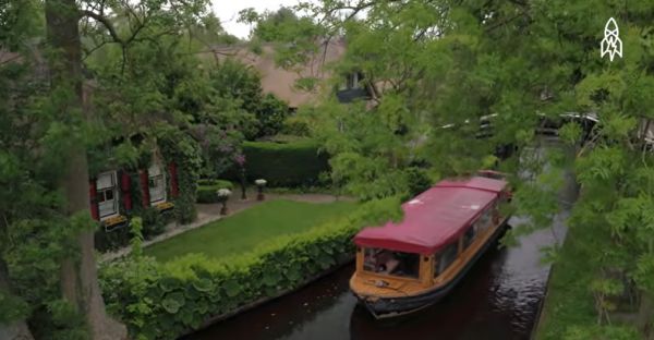 Boats with tourists on a narrow canal, with people walking on pathways and over a small bridge.