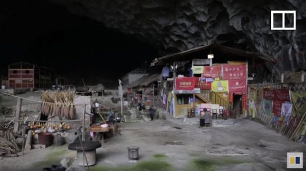 Man standing in a very large cave, with small buildings behind him, and the large entrance to the cave behind the buildings