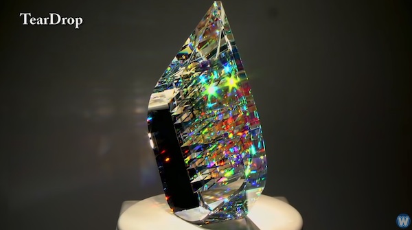 On the left, a cubic piece of glass art with all sorts of colors emanating from within it. On the right, a man holding something similar, with the focus on the glass, and his face in the background blurred.