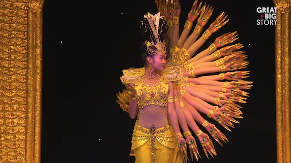 Dancers on stage, standing behind each other so that only the face of the first is visible, but you see a dozen arms and hands in a half-circle pattern to the right of the visible person.