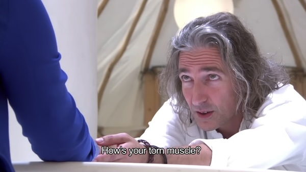 Man with long hair leans over a table looking at a person just off camera in the foreground. He is holding the person's hand, and the caption says, 