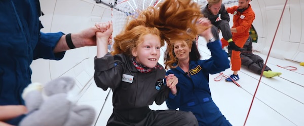 Several people are in an airplane, floating due to very low gravity. In the front is a smiling young girl with long red hair.