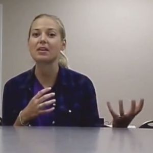 A woman sits behind a table, hands moving as if she's explaining something