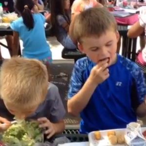 A boy in a blue shirt puts food in his mouth while another boy bends over a bowl of salad to eat