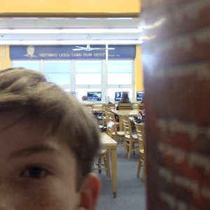 Half of a kid's face is seen from the perspective of a book on a shelf in a crowded library.