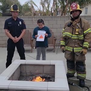 A person holding a drawing of a red triangle stands between two firefighters in front of an outdoor firepit
